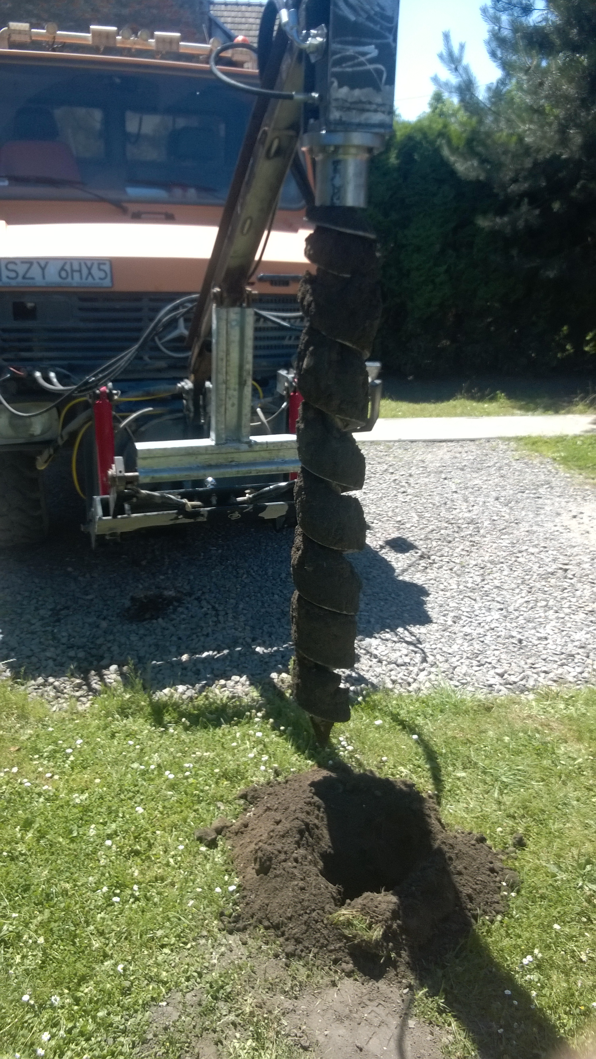 Wiertnica z wiertłem spiralnym, zagłębiona w ziemi na trawniku, z pomarańczowym ciągnikiem w tle. Widoczny gruz przy trawniku.