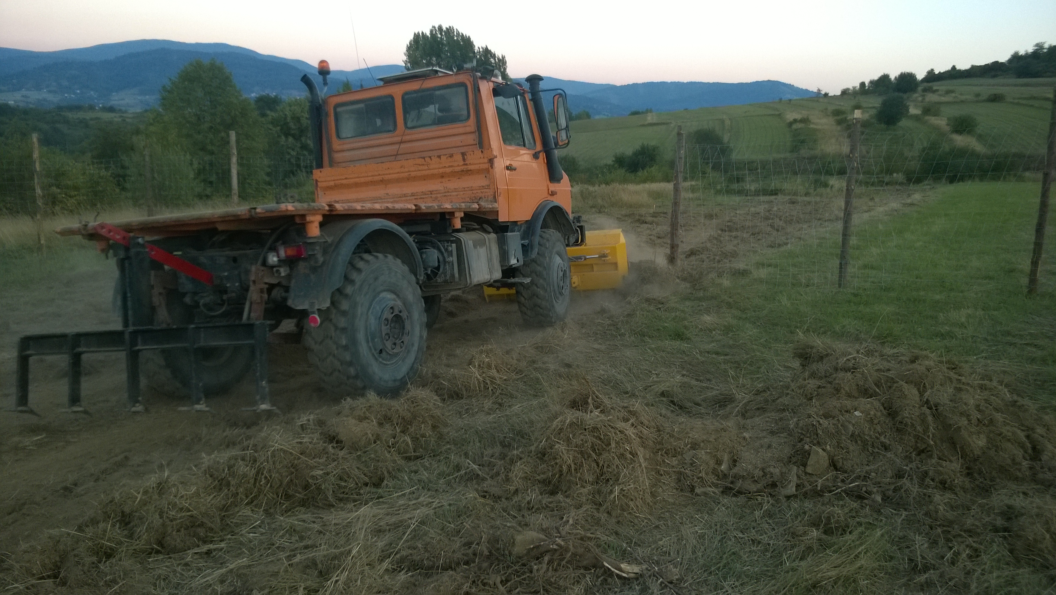 Pomarańczowy Unimog z glebogryzarką w trakcie pracy na polu. W tle widoczne wzgórza i drzewa. Maszyna w ruchu, unoszący się kurz.
