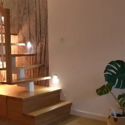 Ażurowe schody z jasnego drewna, zintegrowane z niską zabudową meblową, oświetlone punktowymi lampami, na tle ściany z tapetą w motywy roślinne i zielonego kwiatu doniczkowego.