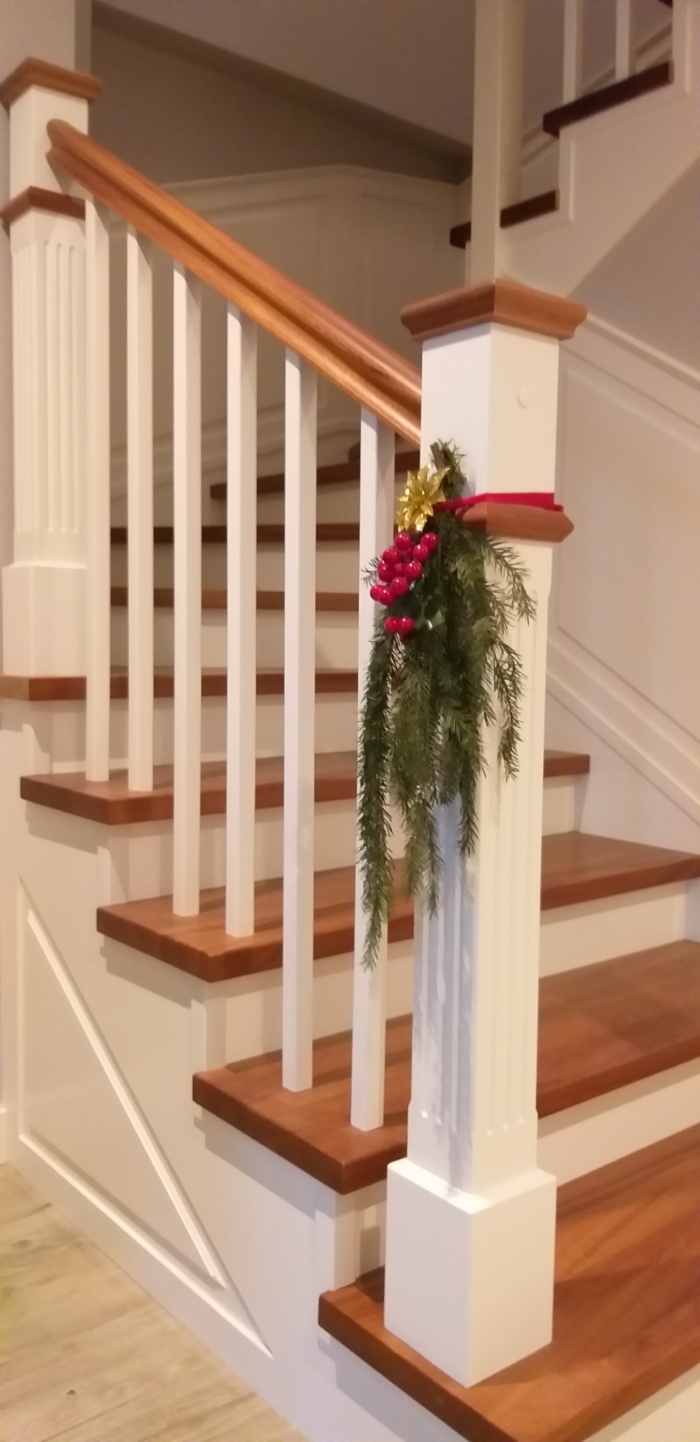 Klasyczne schody z drewnianymi stopniami i białymi balustradami, udekorowane świąteczną girlandą z czerwonymi owocami i złotą kokardą.