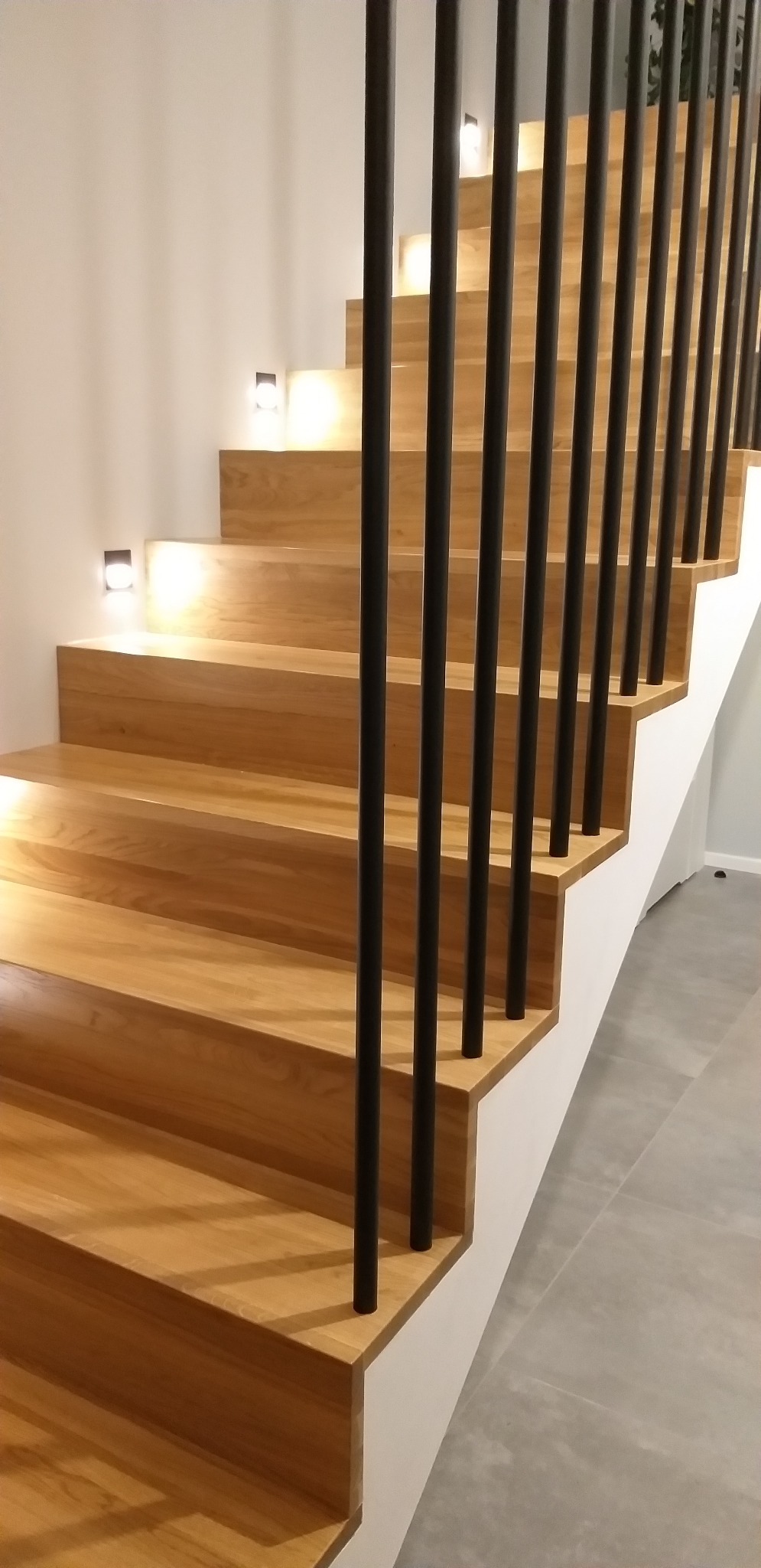 Nowoczesne, oświetlone schody z jasnego drewna z czarnymi, pionowymi balustradami, minimalistyczne oświetlenie punktowe na ścianie.