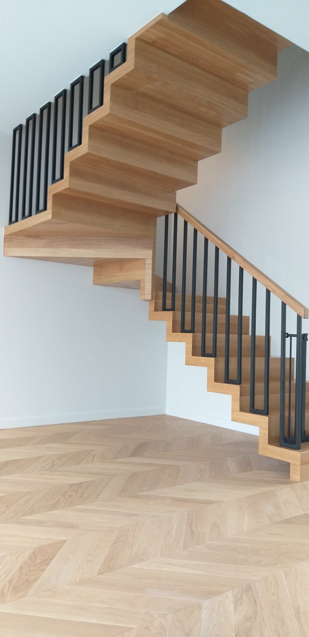 Nowoczesne schody z jasnego drewna z czarnymi, minimalistycznymi balustradami, widok z dołu na parkiet w jodełkę.