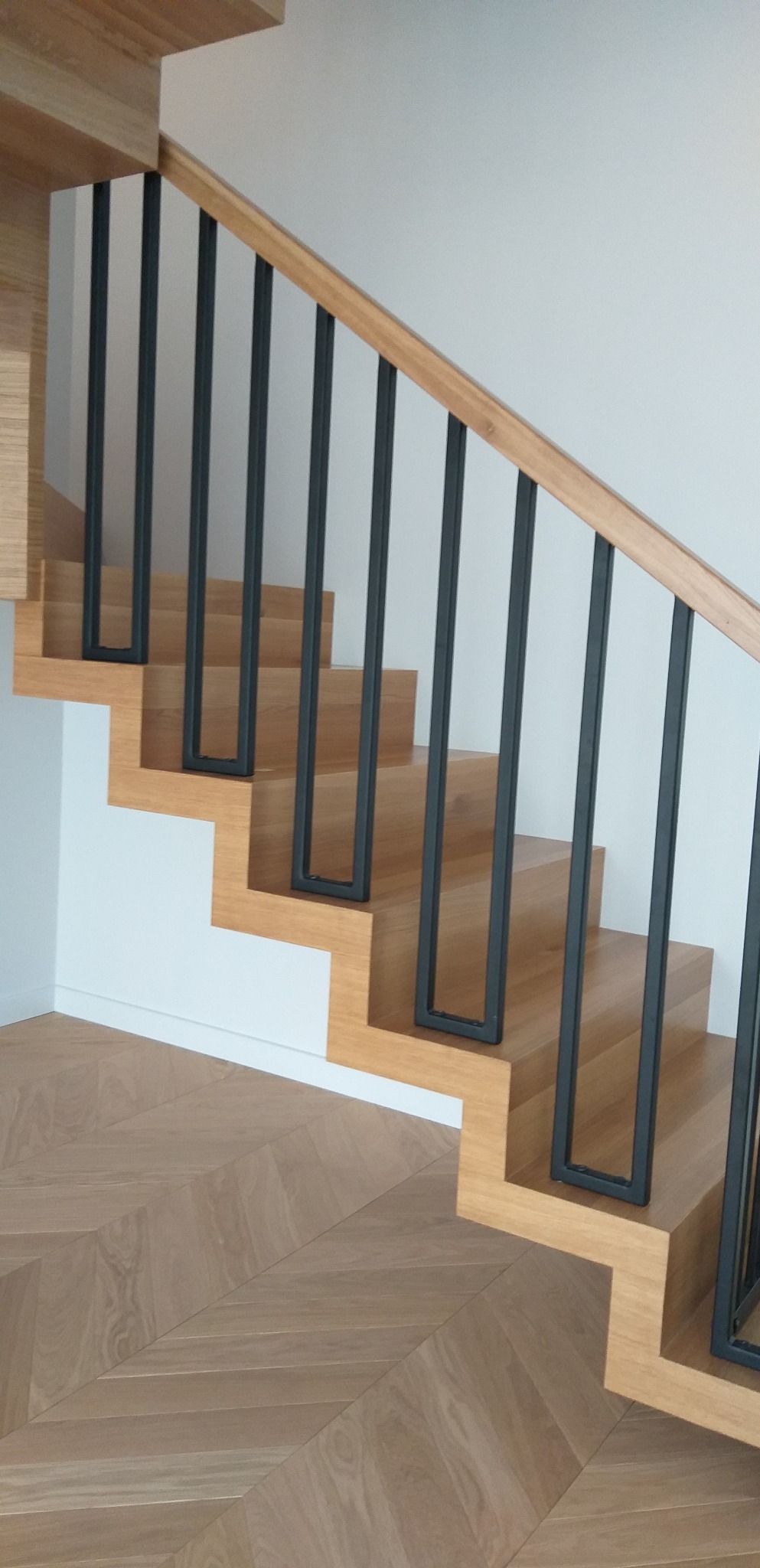 Nowoczesne schody drewniane z czarnymi, prostokątnymi balustradami, parkiet ułożony w jodełkę.