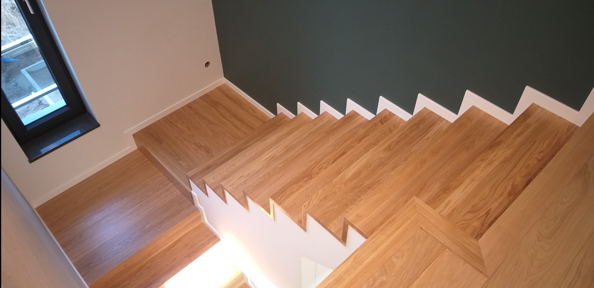 Widok z góry na jasne, dębowe stopnie schodów z białymi podstopnicami, kontrastujące z ciemnozieloną ścianą i fragmentem okna.