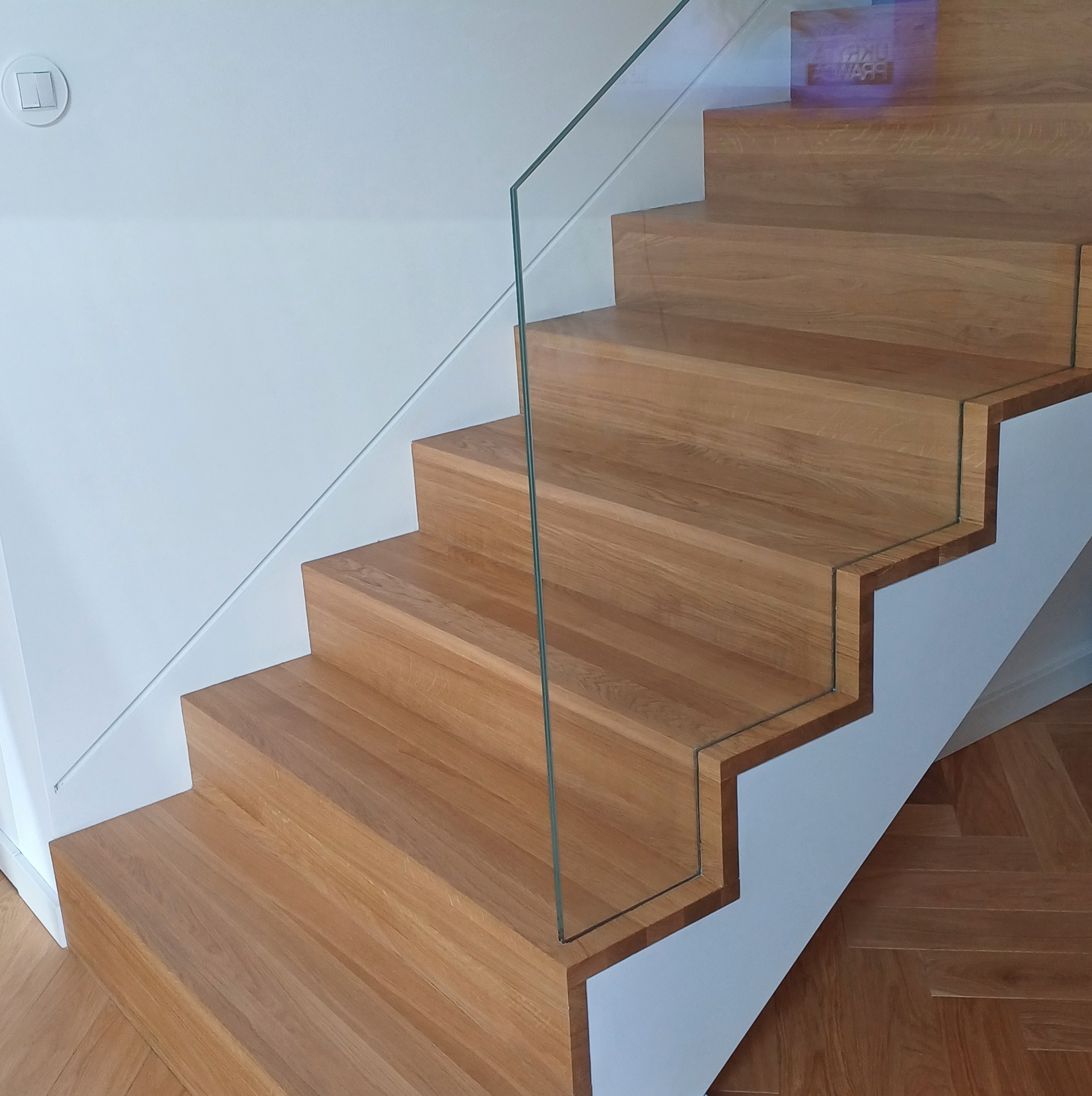 Nowoczesne schody z drewna z balustradą ze szkła, widok z dołu, parkiet w jodełkę na podłodze.