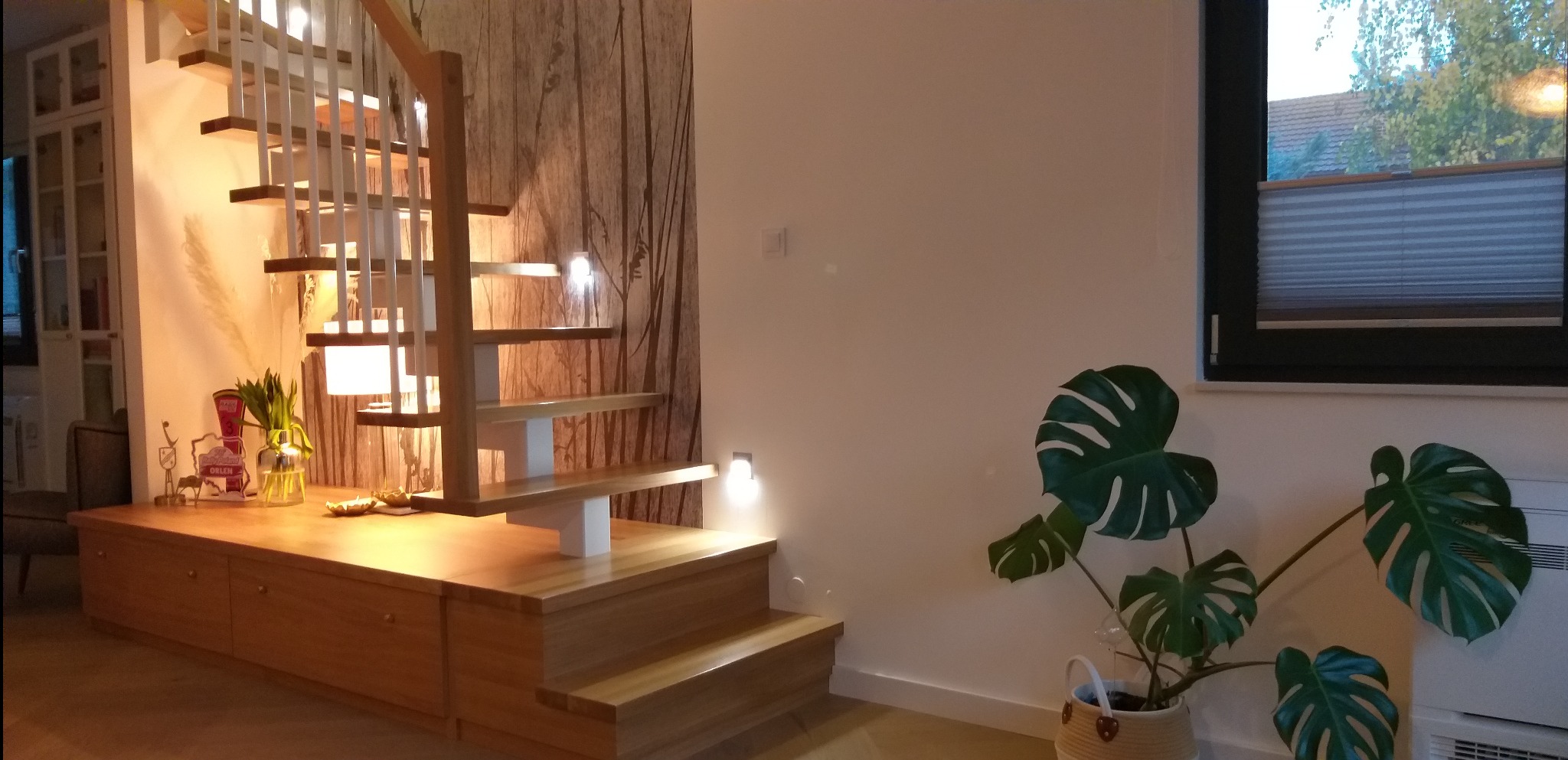 Ażurowe schody z jasnego drewna, zintegrowane z niską zabudową meblową, oświetlone punktowymi lampami, na tle ściany z tapetą w motywy roślinne i zielonego kwiatu doniczkowego.