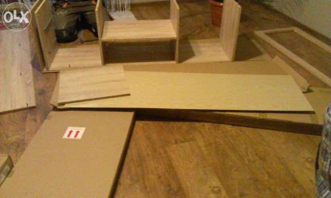 Elementy drewniane, prawdopodobnie do budowy konstrukcji, leżące na podłodze. Widoczne płyty, skrzynki i kartonowe opakowanie z symbolem ostrzegawczym.