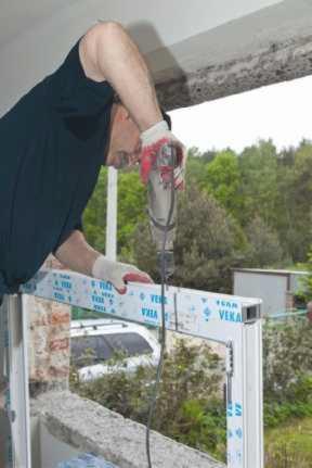 Montaż okna balkonowego VEKA: mężczyzna wierci w profilu, widok na zieleń. Praca z użyciem wiertarki, rękawice ochronne, częściowo widoczny samochód w tle.