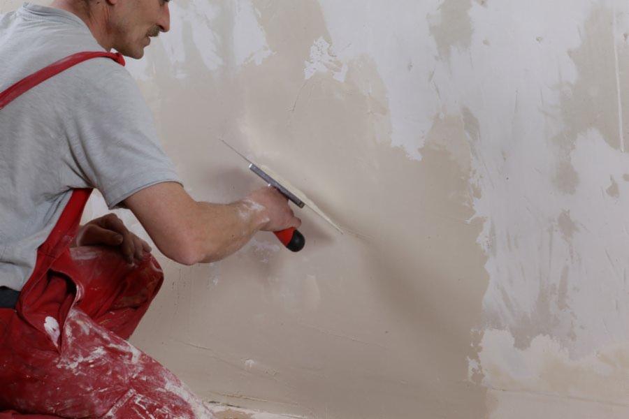 Mężczyzna w czerwonym kombinezonie szpachluje ścianę szpachelką. Widoczny fragment ściany z nałożoną gładzią. Praca wykończeniowa w trakcie postępu.