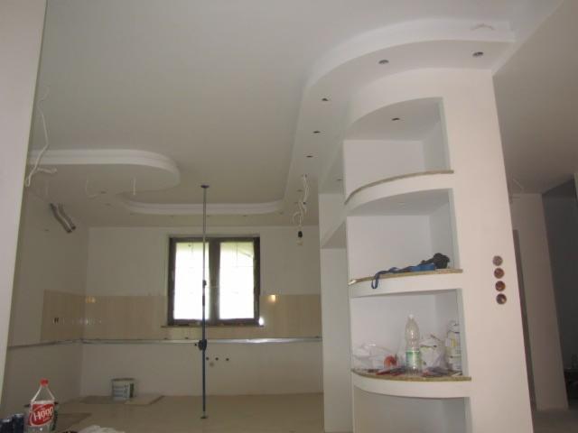 Wnętrze w trakcie prac wykończeniowych: dekoracyjny sufit podwieszany z oświetleniem punktowym, kolumna z półkami. Widoczne niedokończone instalacje elektryczne.