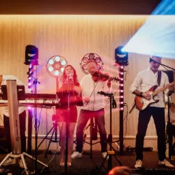 Zespół muzyczny w akcji: wokalistka z mikrofonem, skrzypek, gitarzysta i klawiszowiec. Oświetlenie sceniczne i dekoracyjne tło z fakturą.