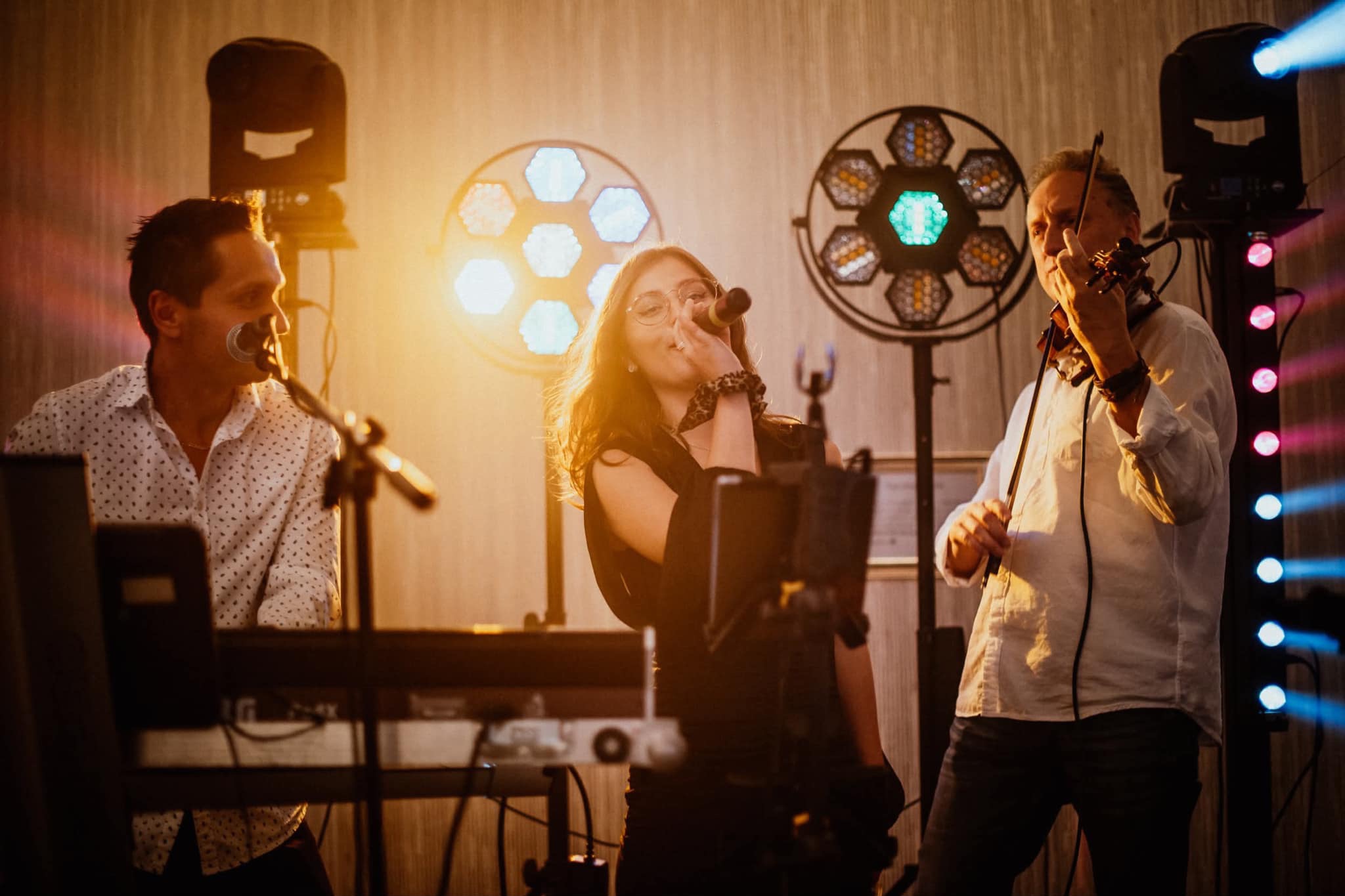 Zespół muzyczny na scenie: wokalistka z mikrofonem, klawiszowiec i skrzypek, oświetlenie sceniczne. Ciepłe, dynamiczne ujęcie z występu na żywo.