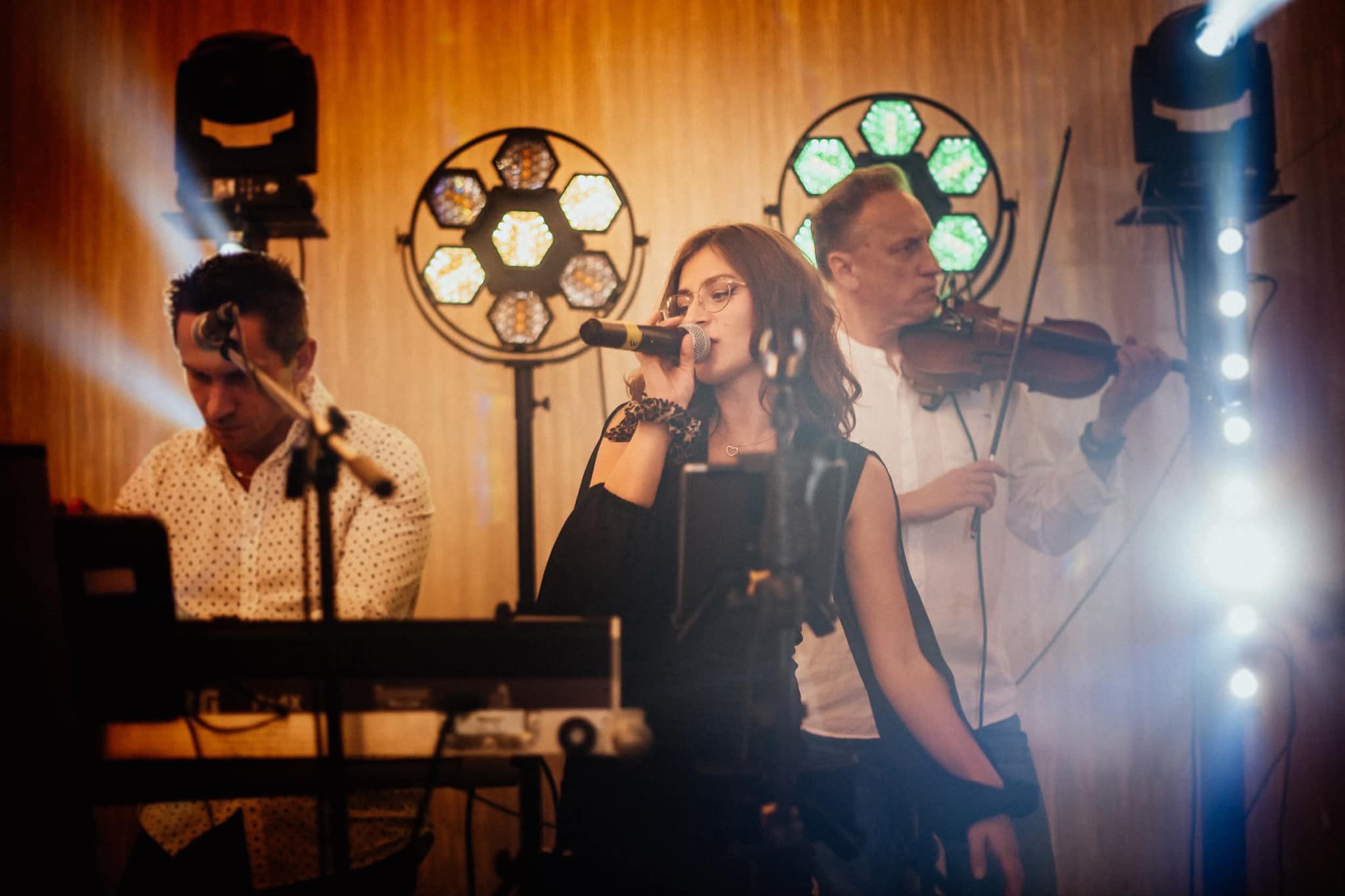 Zespół muzyczny na scenie: wokalistka z mikrofonem, skrzypek i klawiszowiec, oświetlenie sceniczne. Koncert w trakcie występu.
