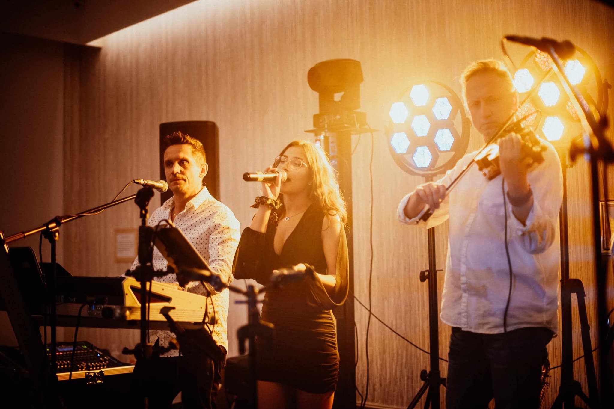 Zespół muzyczny w akcji: klawiszowiec, wokalistka i skrzypek, oświetleni reflektorami punktowymi, tworzą energetyczny nastrój na scenie.