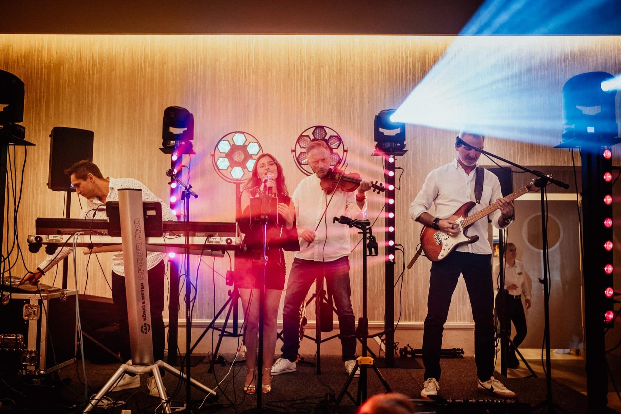 Zespół muzyczny w akcji: wokalistka z mikrofonem, skrzypek, gitarzysta i klawiszowiec. Oświetlenie sceniczne i dekoracyjne tło z fakturą.