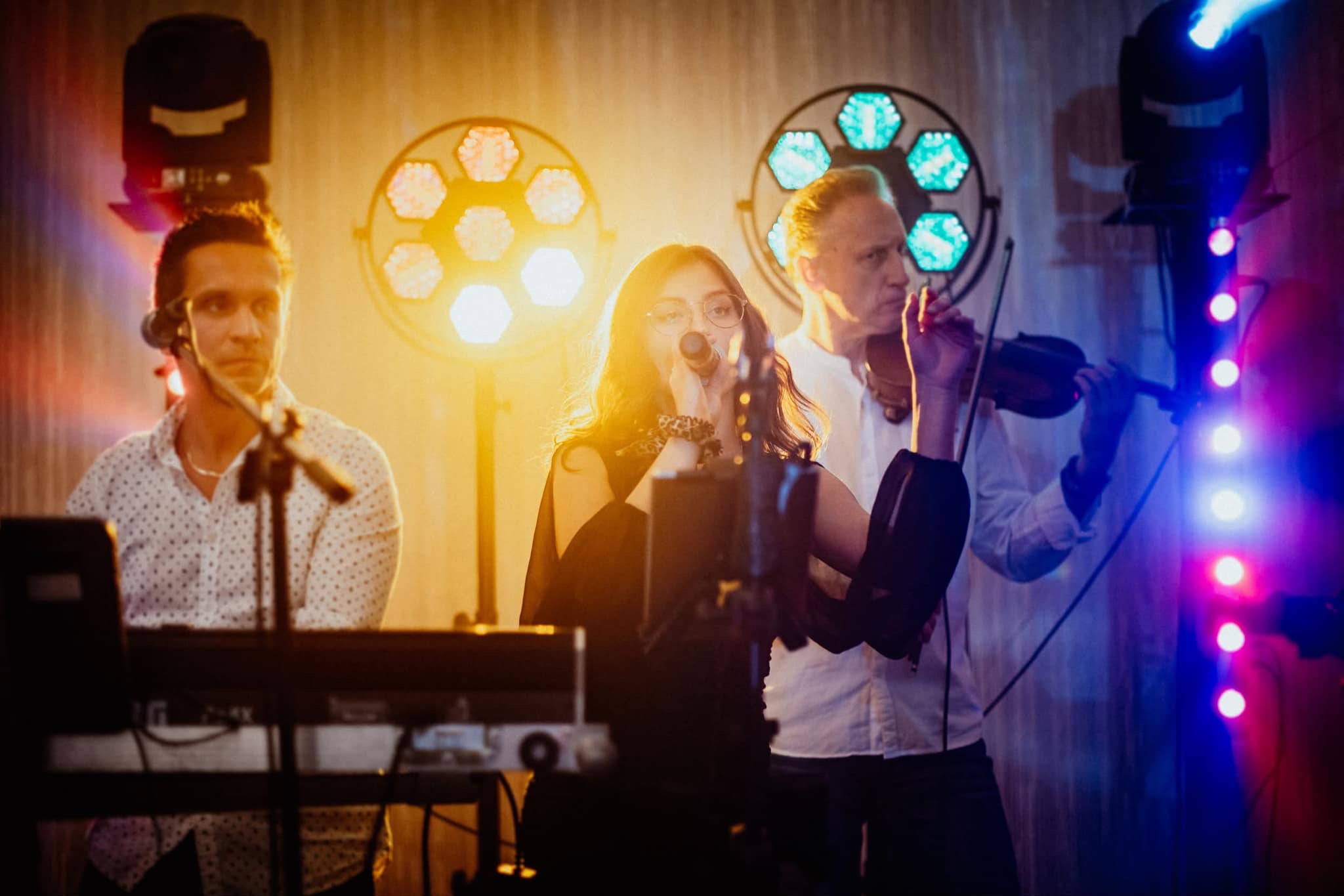 Zespół muzyczny w akcji: wokalistka śpiewa do mikrofonu, skrzypek gra, a klawiszowiec akompaniuje, wszystko w blasku kolorowych świateł scenicznych.