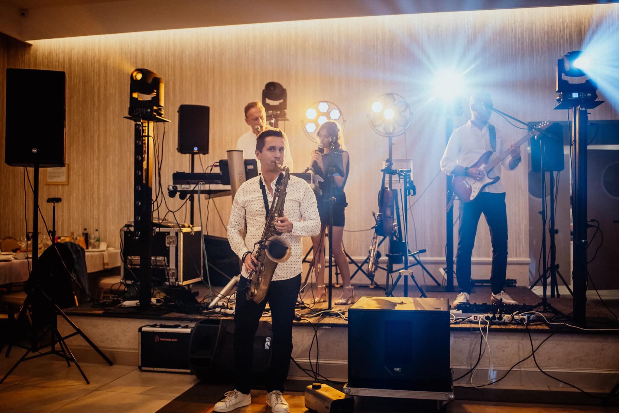 Zespół muzyczny na scenie: saksofonista na pierwszym planie, w tle wokalistka, gitarzysta i klawiszowiec. Widoczne nagłośnienie i oświetlenie sceniczne.