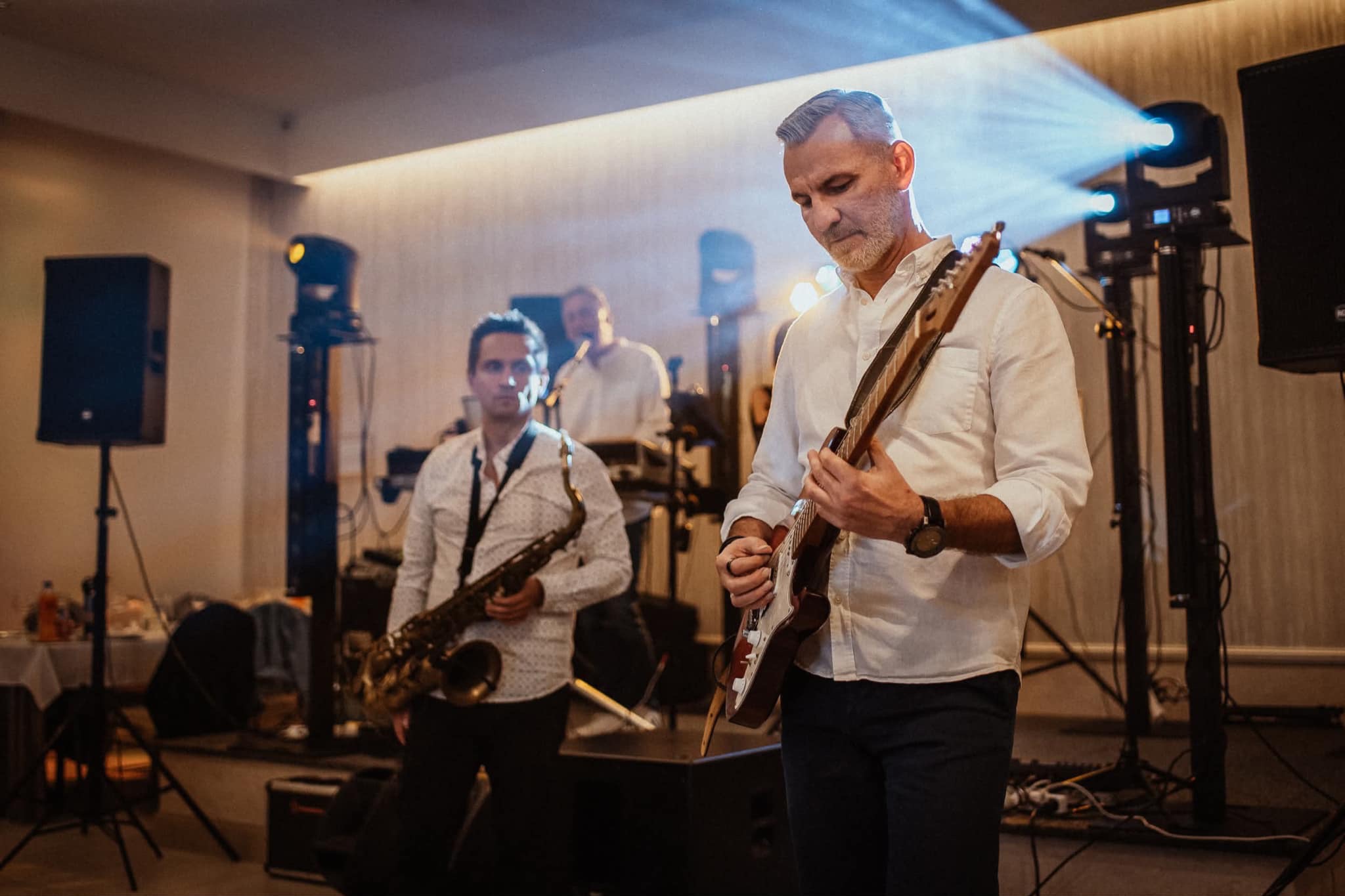 Zespół muzyczny na scenie: gitarzysta skupiony na grze, saksofonista w tle i perkusista. Oświetlenie sceniczne tworzy atmosferę koncertu. Białe koszule muzyków.