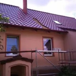 Ocieplony dom z nowym, brązowym dachem i oknami PCV. Widoczne rusztowanie, fragment drzewa i mała dobudówka z łukowym wejściem. Słoneczny dzień.