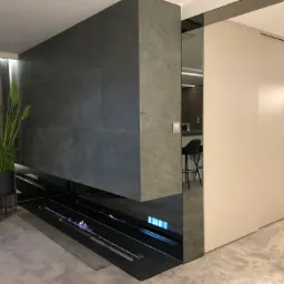 Nowoczesna ściana z kamienia z wbudowanym biokominkiem, odbicie kuchni w szklanej powierzchni, minimalistyczny design wnętrza.