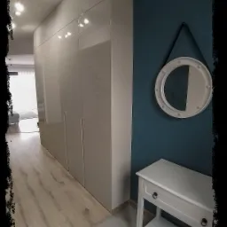 Długa szafa wnękowa z połyskującymi, beżowymi frontami, zlicowana ze ścianą w przedpokoju, obok biała konsola z okrągłym lustrem na turkusowej ścianie.