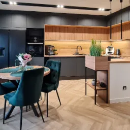 Nowoczesna kuchnia z ciemnymi, matowymi frontami szafek i drewnianymi akcentami, widoczny stół z zielonymi krzesłami i parkiet w jodełkę.