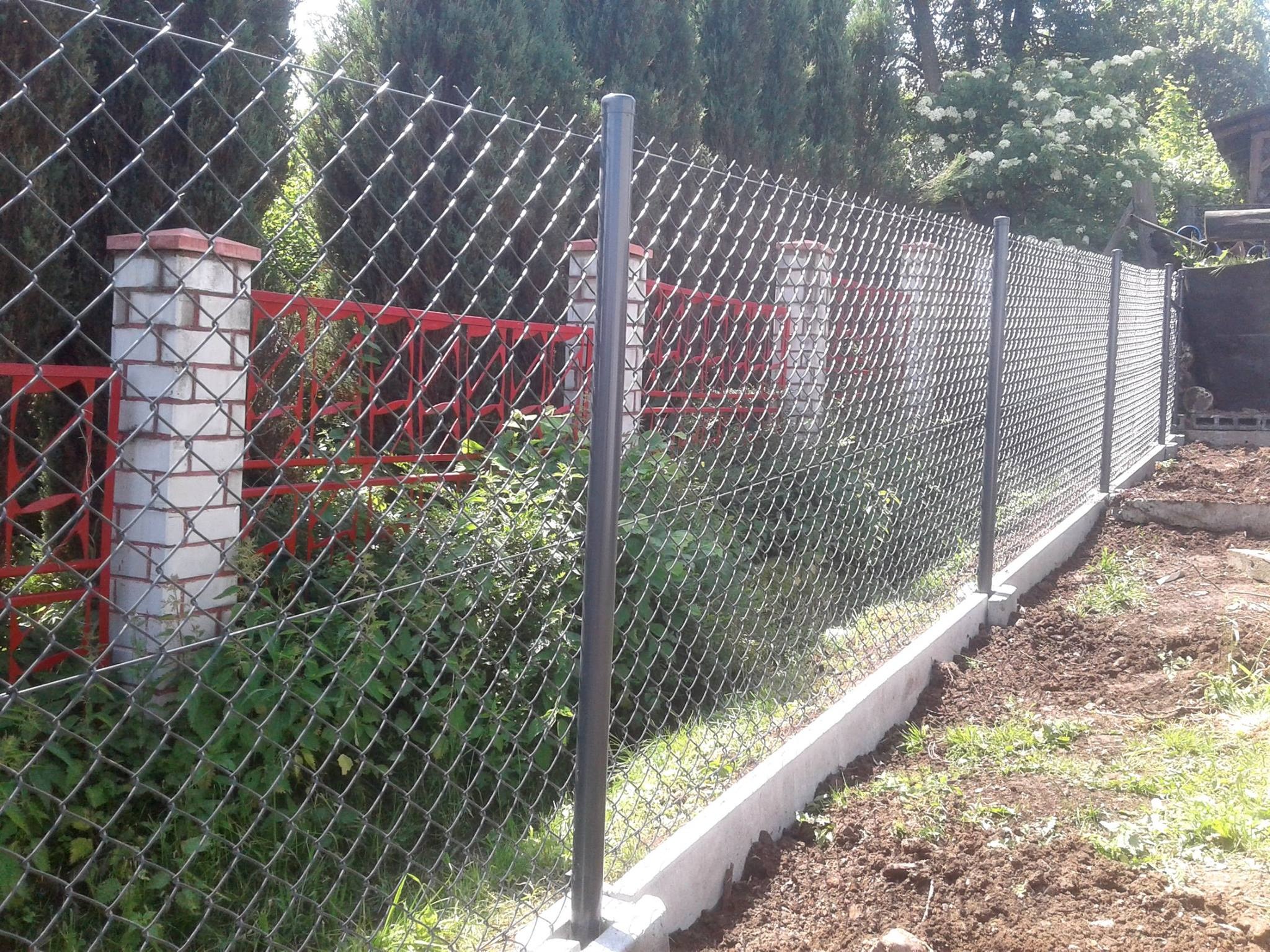 Nowo zainstalowane ogrodzenie z siatki z betonową podmurówką i szarymi słupkami, częściowo zasłonięte roślinnością, z widocznymi elementami czerwonego ogrodzenia w tle.