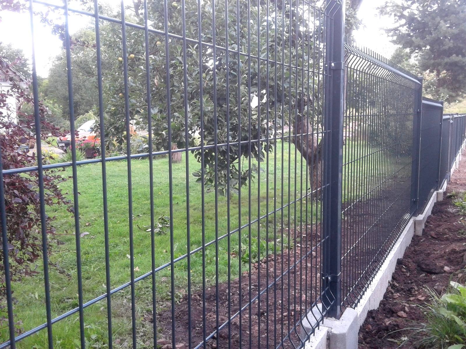 Nowo zamontowane, ciemne ogrodzenie panelowe z podmurówką betonową na tle zielonego trawnika i drzew z owocami.