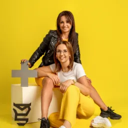 Dwie uśmiechnięte kobiety w żółtych stylizacjach pozują na żółtym tle, trzymając szary znak plusa i siedząc obok logotypu firmy.