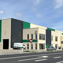 ArchiDOMO Biuro Projektów - Nowoczesna hala magazynowa z elementami zieleni i szarości, widoczna z perspektywy ulicy. Na pierwszym planie droga z oznakowaniem poziomym i kilka samochodów.