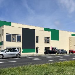 ArchiDOMO Biuro Projektów - Nowoczesna hala magazynowa z elementami zieleni i szarości, zaparkowane samochody, błękitne niebo. Widok z poziomu ulicy, zieleń w tle.