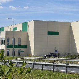 ArchiDOMO Biuro Projektów - Nowoczesna hala magazynowa z elementami zieleni, widoczna zza barierek. Tablica z nazwą miejscowości 'Rzegnowo 1,5'. Słoneczny dzień, błękitne niebo z chmurami.