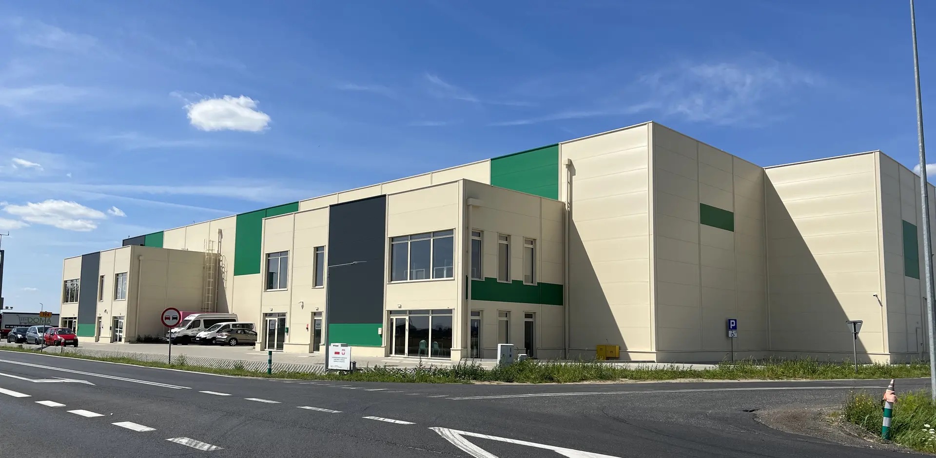 Nowoczesny budynek usługowy z elementami zieleni i szarości, widoczny z perspektywy ulicy w słoneczny dzień. Na pierwszym planie fragment jezdni.