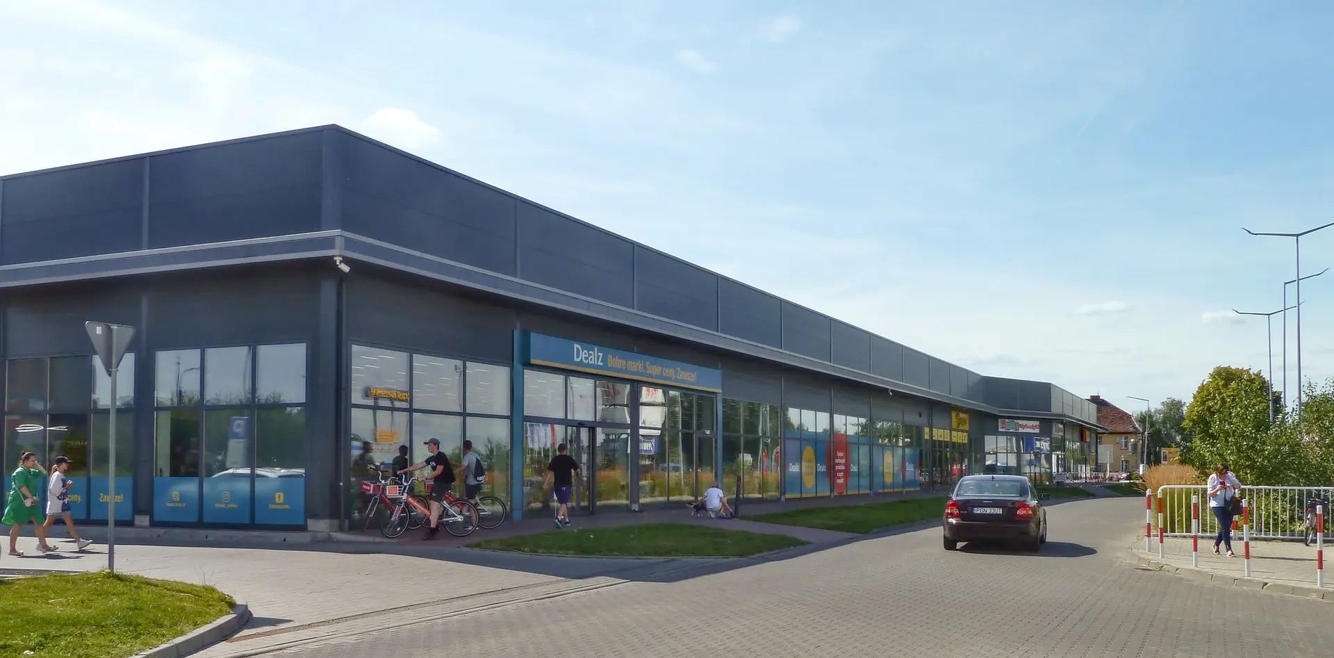 Nowoczesny budynek usługowy z szarym wykończeniem, przeszklonymi witrynami sklepów Dealz i Media Expert, z widoczną kostką brukową i przechodniami w słoneczny dzień.