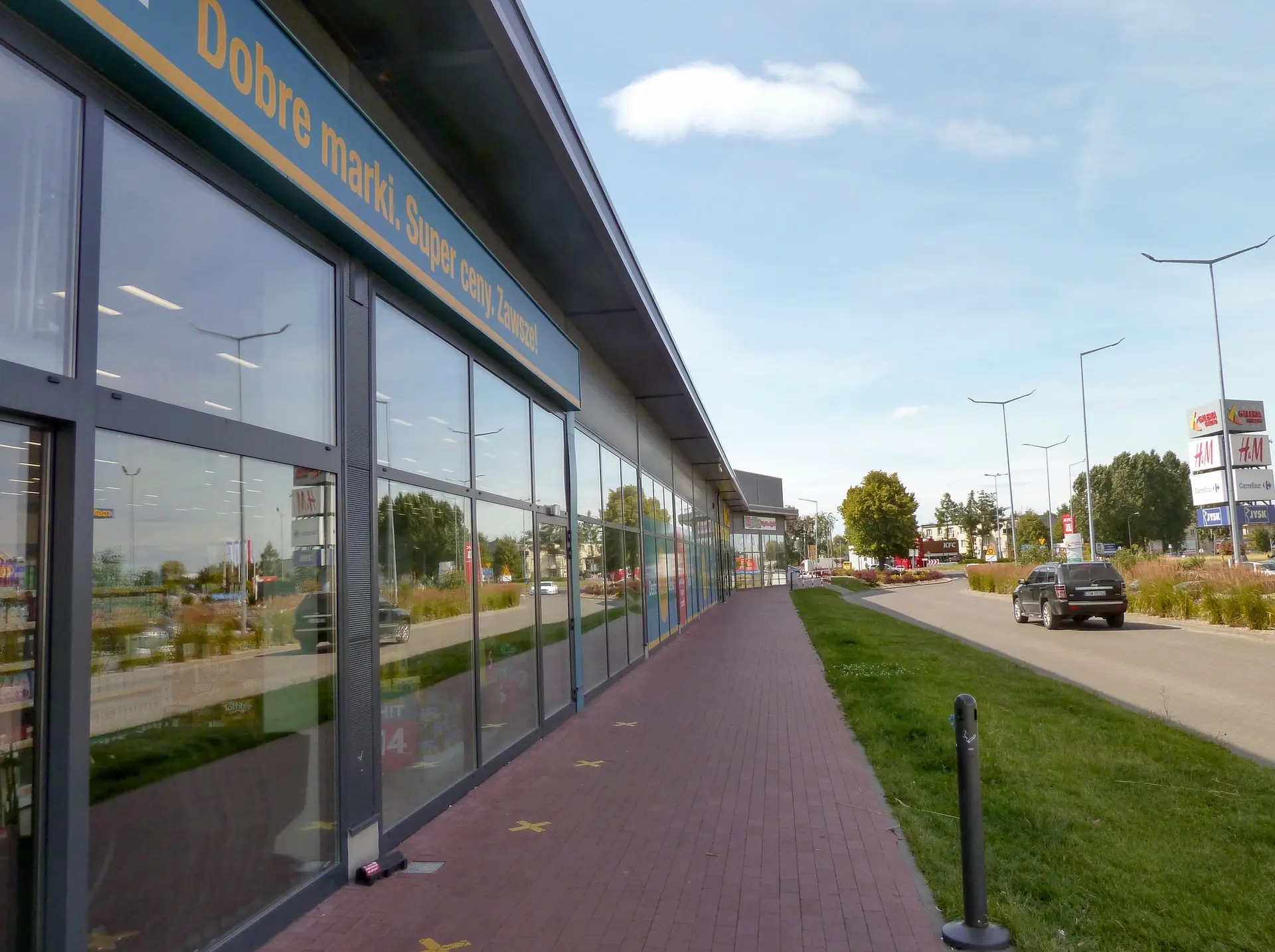 Nowoczesny budynek usługowy z dużymi przeszkleniami i napisem reklamowym, z widocznym parkingiem i znakiem drogowym w tle.