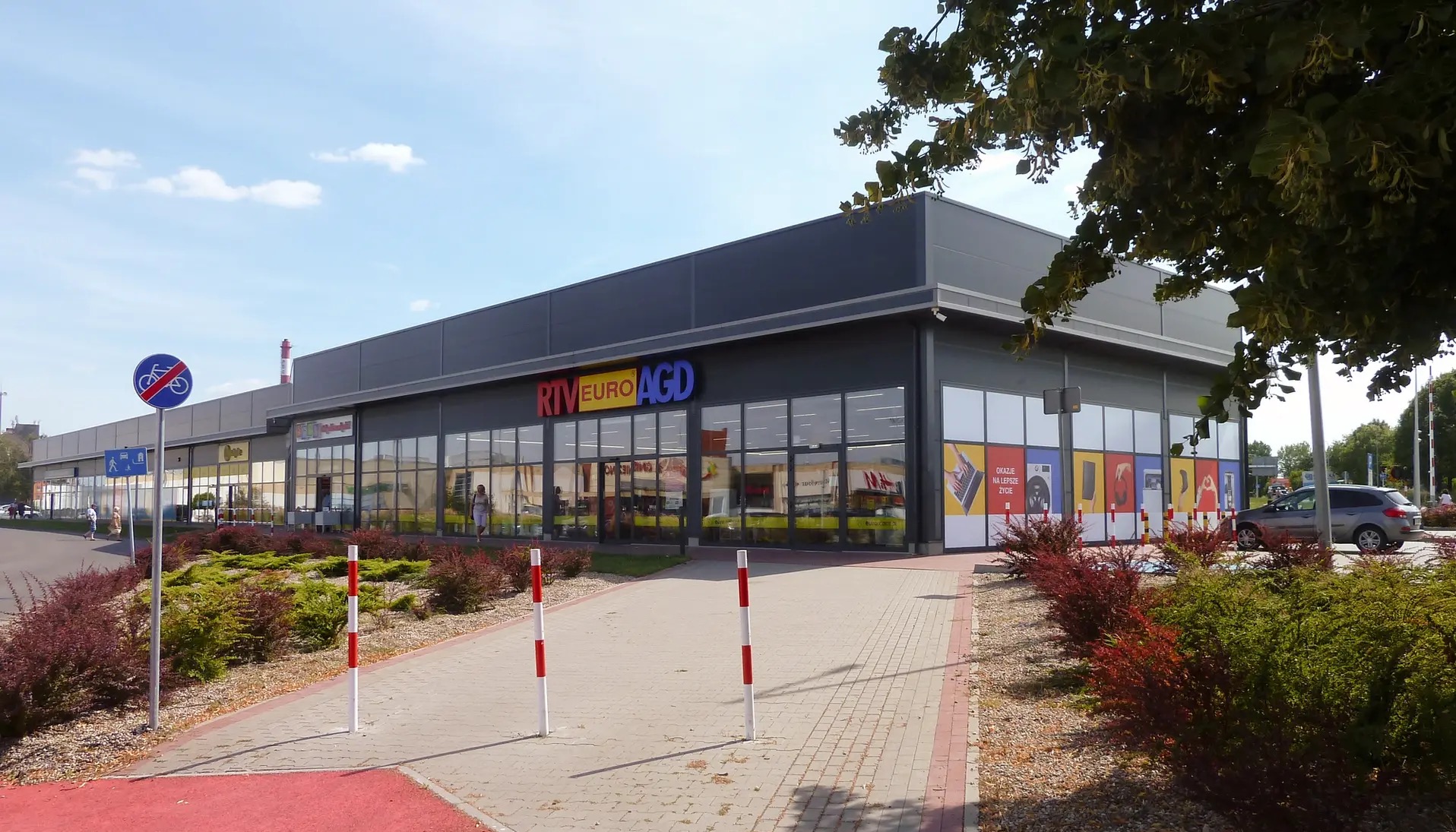 Nowoczesny budynek usługowy RTV Euro AGD z szarą elewacją, dużymi oknami i kolorowym logo, otoczony zielenią i chodnikiem z barierkami.