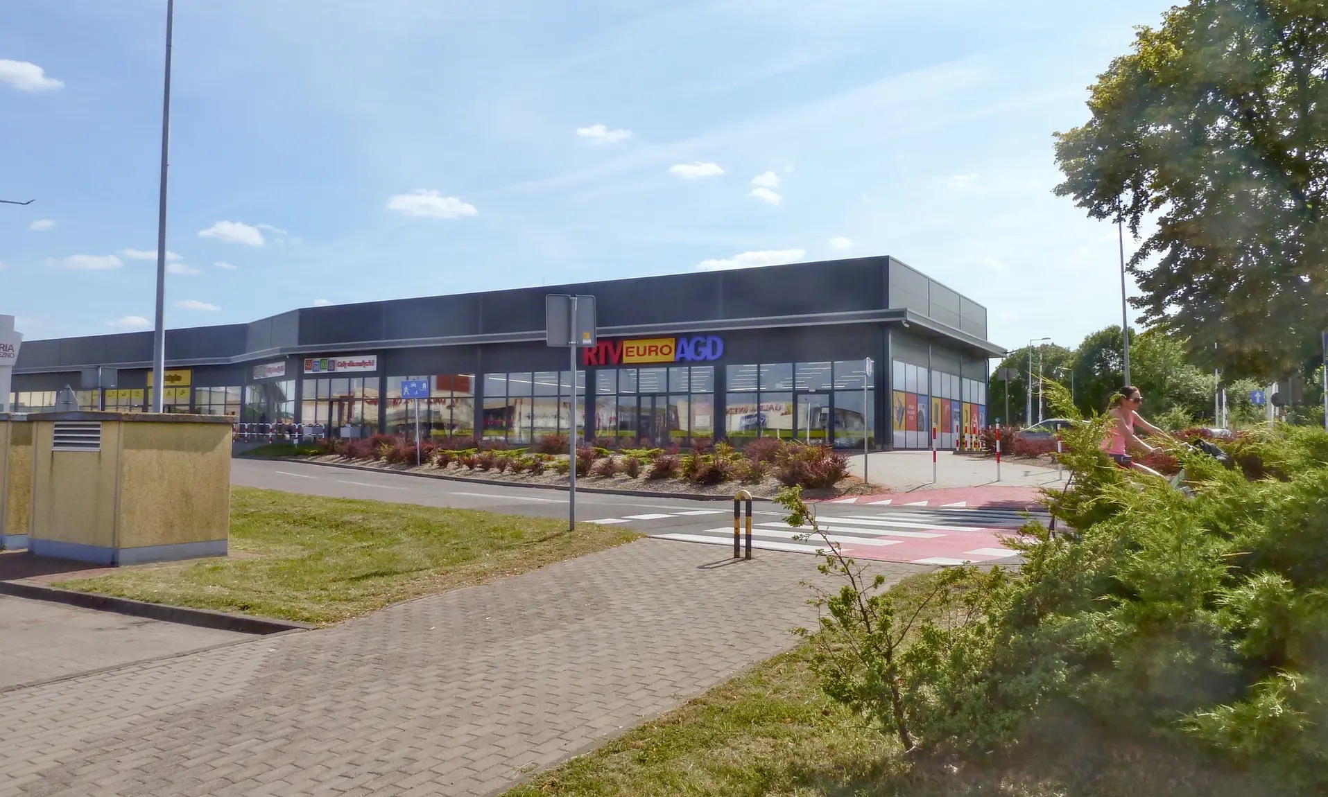 Nowoczesny budynek usługowy z dużymi oknami i logo RTV Euro AGD, widziany z poziomu chodnika w słoneczny dzień. Widoczny fragment przejścia dla pieszych.