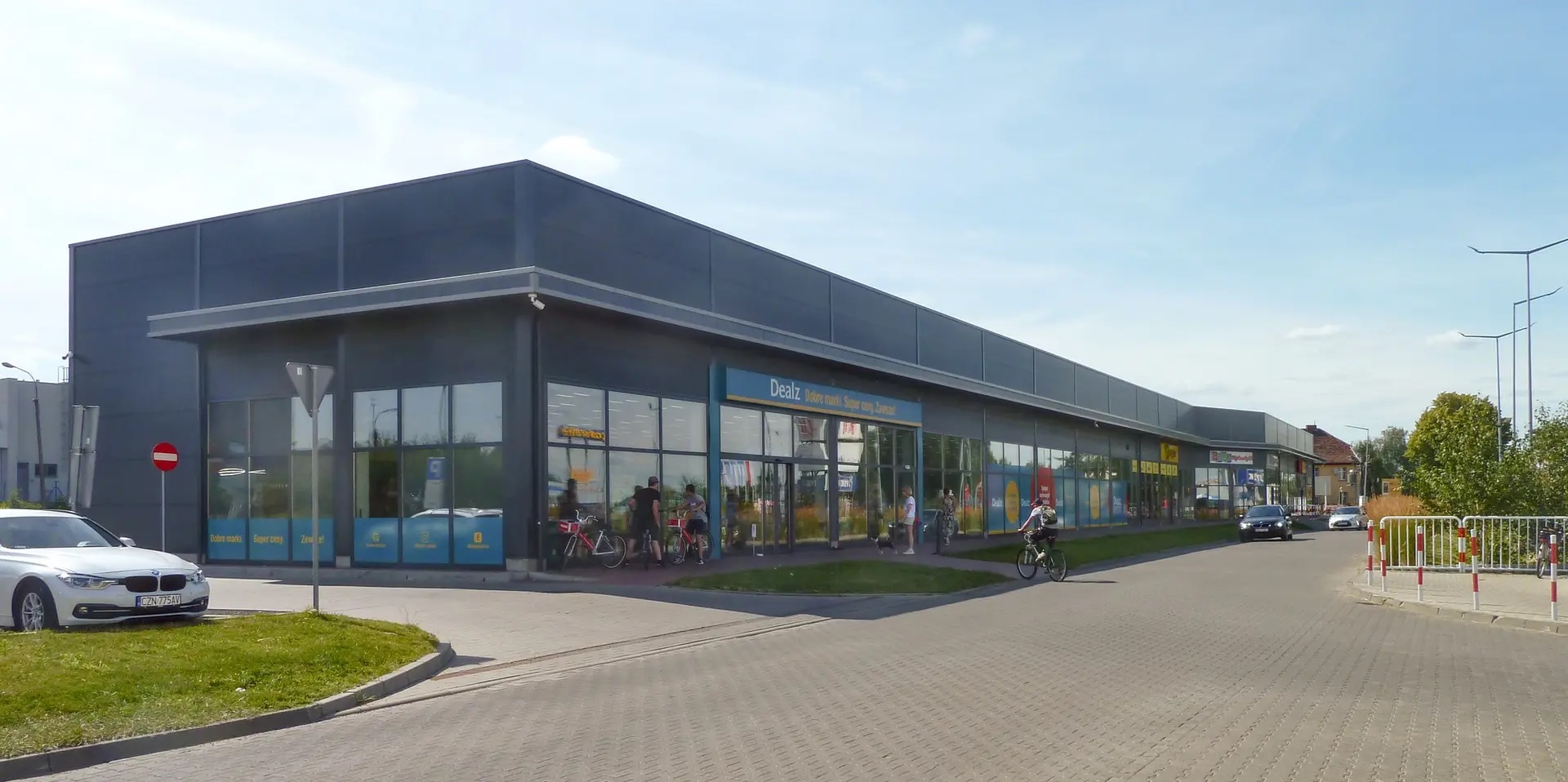 Nowoczesny budynek usługowy z szarą elewacją i dużymi oknami, przed którym znajdują się osoby i zaparkowany samochód. Widoczny znak zakazu wjazdu.