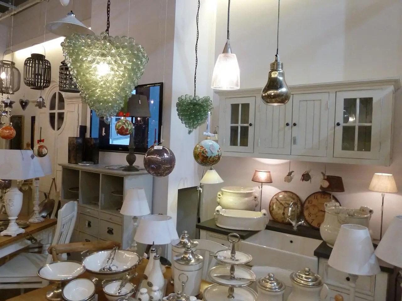 Wnętrze sklepu z oświetleniem: wiszące lampy o fantazyjnych kształtach, meble w stylu vintage, dekoracyjne naczynia i zegary tworzą eklektyczną kompozycję.