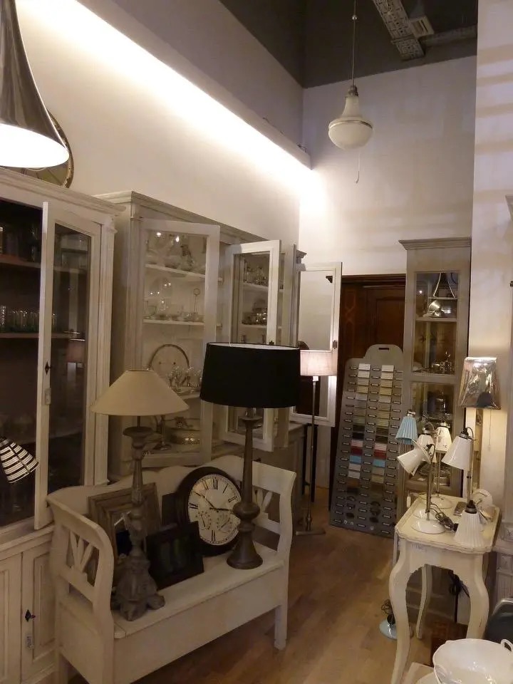 Jasne wnętrze: eklektyczny salon z witrynami, lampami i zegarem na ławce. Neutralna paleta barw, drewniana podłoga, wysokie sufity z oświetleniem.