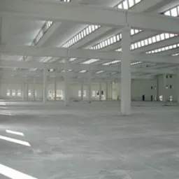 Wnętrze hali przemysłowej w jasnych barwach. Widoczne betonowe filary i posadzka. Naturalne światło wpadające przez okna w dachu. Minimalizm i surowość wykończenia.