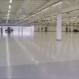 Jasnoszara, gładka posadzka przemysłowa w hali magazynowej z białymi kolumnami i oświetleniem liniowym pod sufitem.