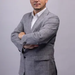 Portret mężczyzny w okularach i szarym garniturze w kratę, z rękami skrzyżowanymi na piersiach, na jasnoszarym tle.