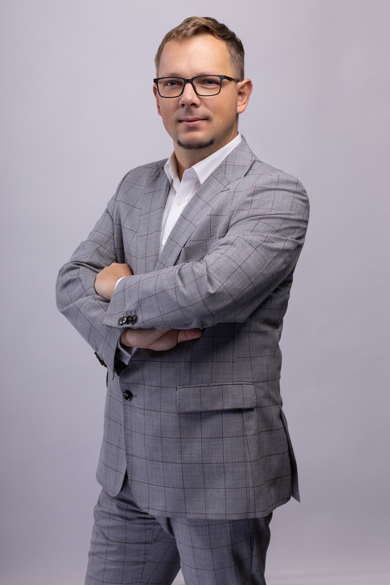 Portret mężczyzny w okularach i szarym garniturze w kratę, z rękami skrzyżowanymi na piersiach, na jasnoszarym tle.