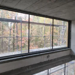 Surowy betonowy strop i ściany z białych bloczków w trakcie budowy domu z widokiem na jesienny las przez duże okno; rusztowania wewnątrz i na zewnątrz.