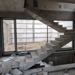 Surowy betonowy szkielet domu w budowie z widocznymi schodami betonowymi, oknami i ścianami z betonu komórkowego. Na podłodze materiały budowlane i drabina.