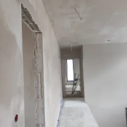 Wnętrze w trakcie tynkowania maszynowego, widoczne surowe ościeżnice drzwiowe, ślady po instalacjach elektrycznych i drabina w tle.