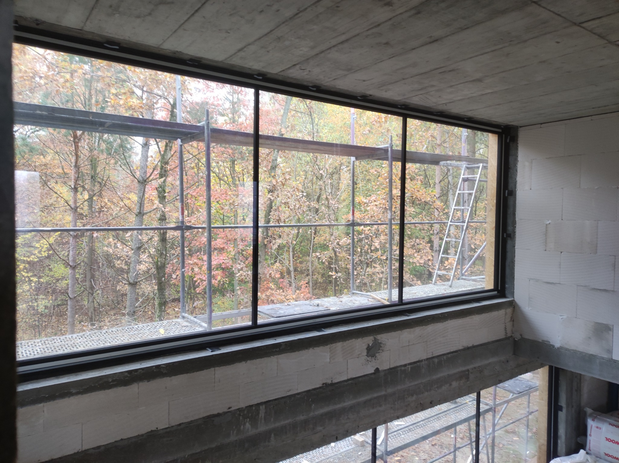 Surowy betonowy strop i ściany z białych bloczków w trakcie budowy domu z widokiem na jesienny las przez duże okno; rusztowania wewnątrz i na zewnątrz.