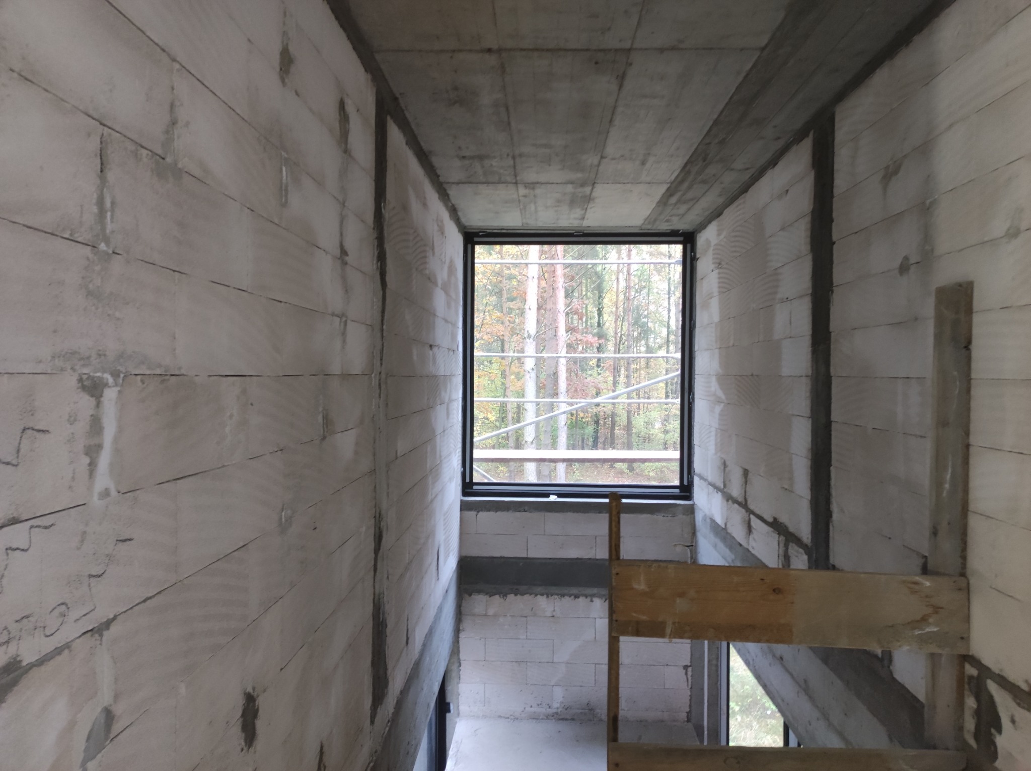 Wnętrze surowego budynku w trakcie budowy, ściany z białych bloczków, betonowy strop, okno z widokiem na jesienny las, fragment drewnianego rusztowania.