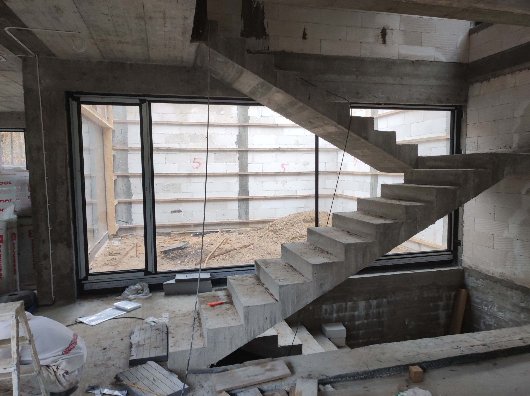 Surowy betonowy szkielet domu w budowie z widocznymi schodami betonowymi, oknami i ścianami z betonu komórkowego. Na podłodze materiały budowlane i drabina.